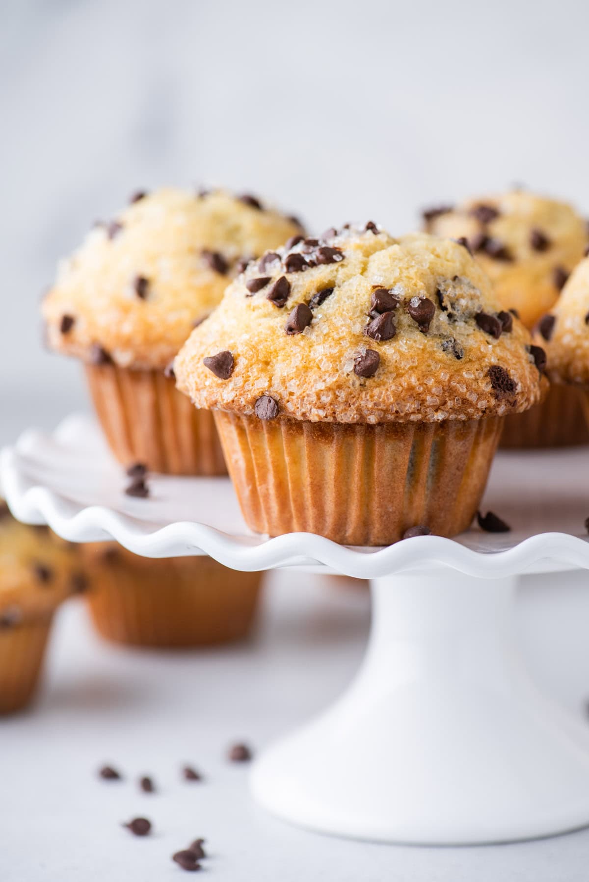 Chocolate chip muffins on a white, ruffled cake stand, with more muffins and scattered chocolate chips in the background on a light surface.