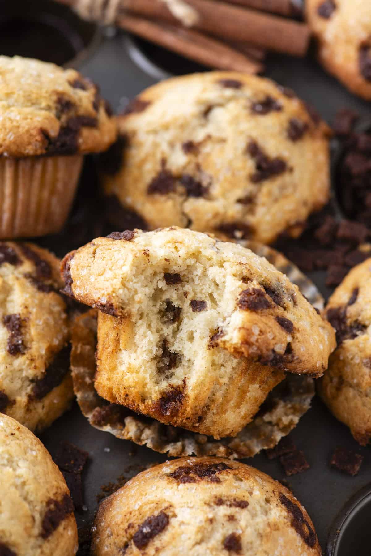 A close-up of several cinnamon chip muffins, with one muffin in the center partially split open to reveal its moist interior and cinnamon chips. Cinnamon sticks and cinnamon pieces are in the background.