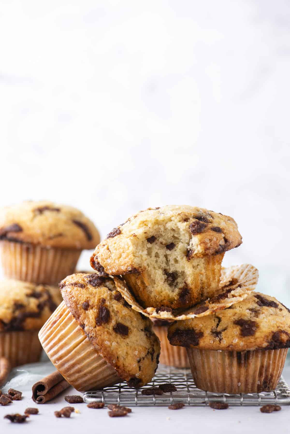 A stack of cinnamon chip muffins sits on a cooling rack, with one muffin cut open to show its soft, moist interior. The background is light and blurred, highlighting the muffins.