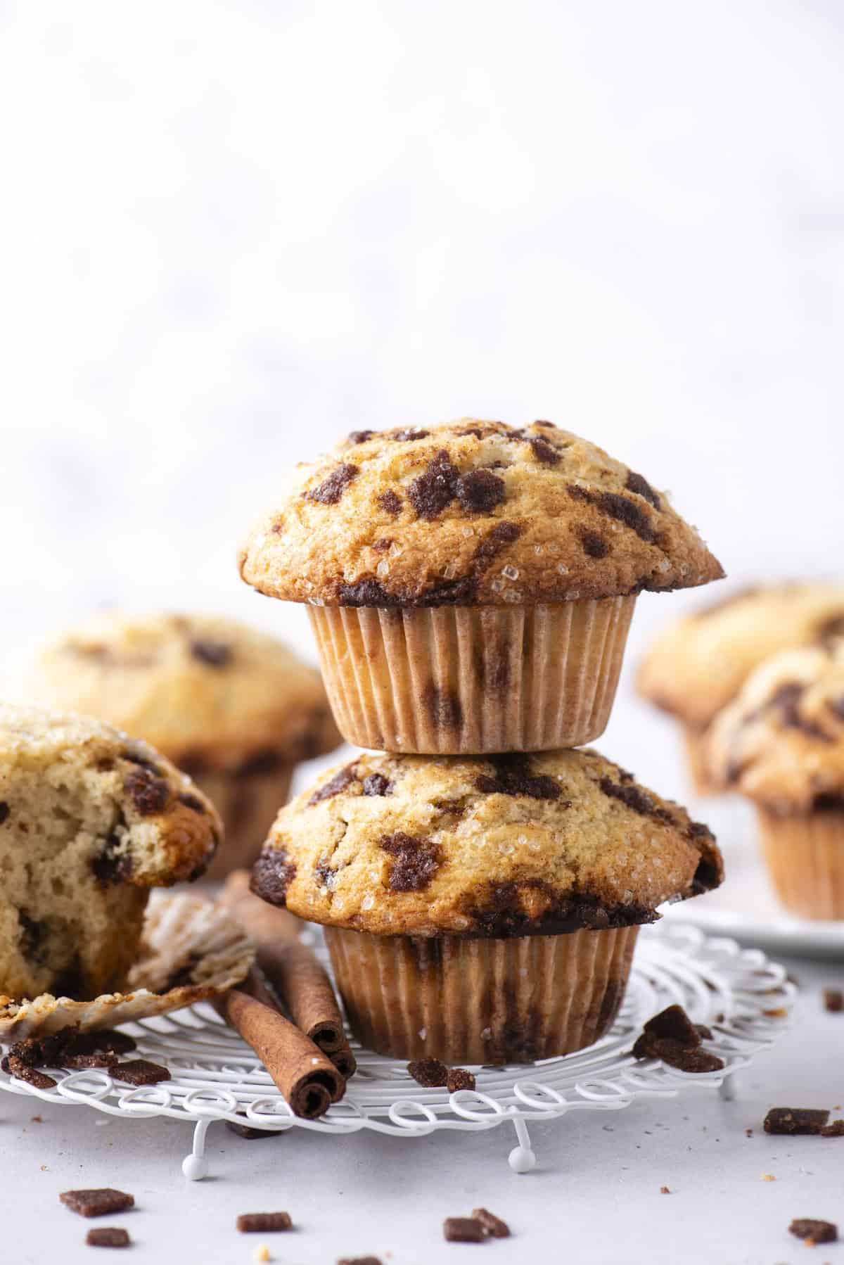 Two cinnamon chip muffins stacked on a white wire rack with additional muffins and loose cinnamon chunks in the background. A partially eaten muffin and cinnamon sticks are also visible.