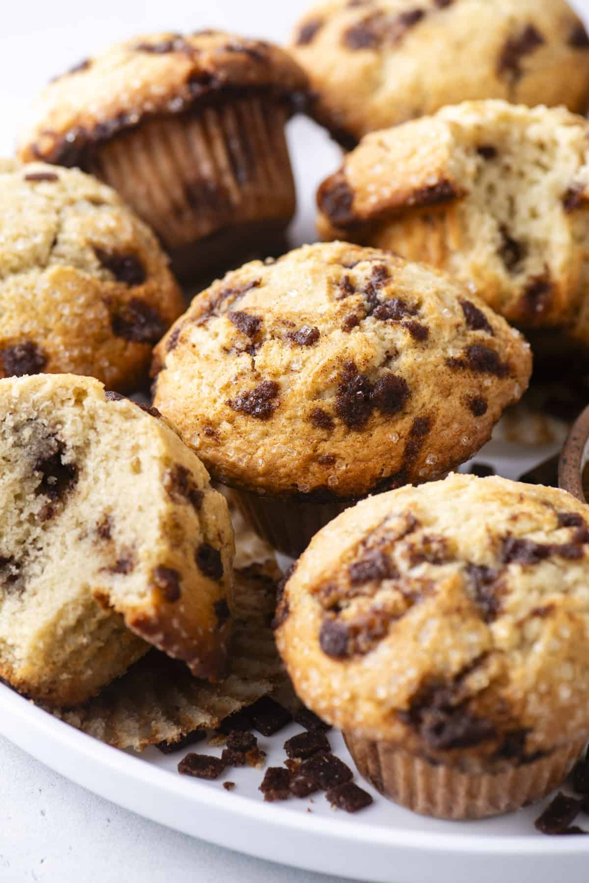 A close-up of several cinnamon chip muffins on a white plate, with one muffin partially broken open to show the moist interior and scattered chocolate chips. Crumbs are visible on the plate.