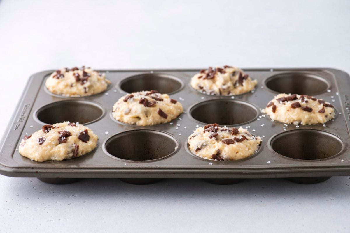 A muffin tin with eight wells, four of which are filled with unbaked muffin batter topped with cinnamon chips and a sprinkle of salt, sits on a light gray surface.