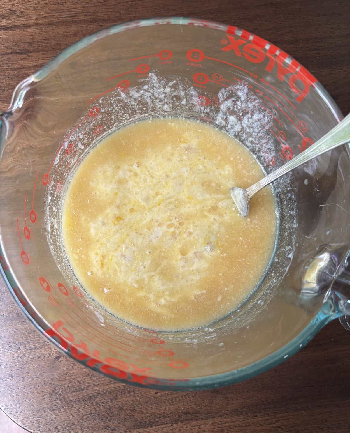 A glass measuring cup with a mixture of wet ingredients and some flour, being mixed with a metal spoon, sits on a wooden surface.