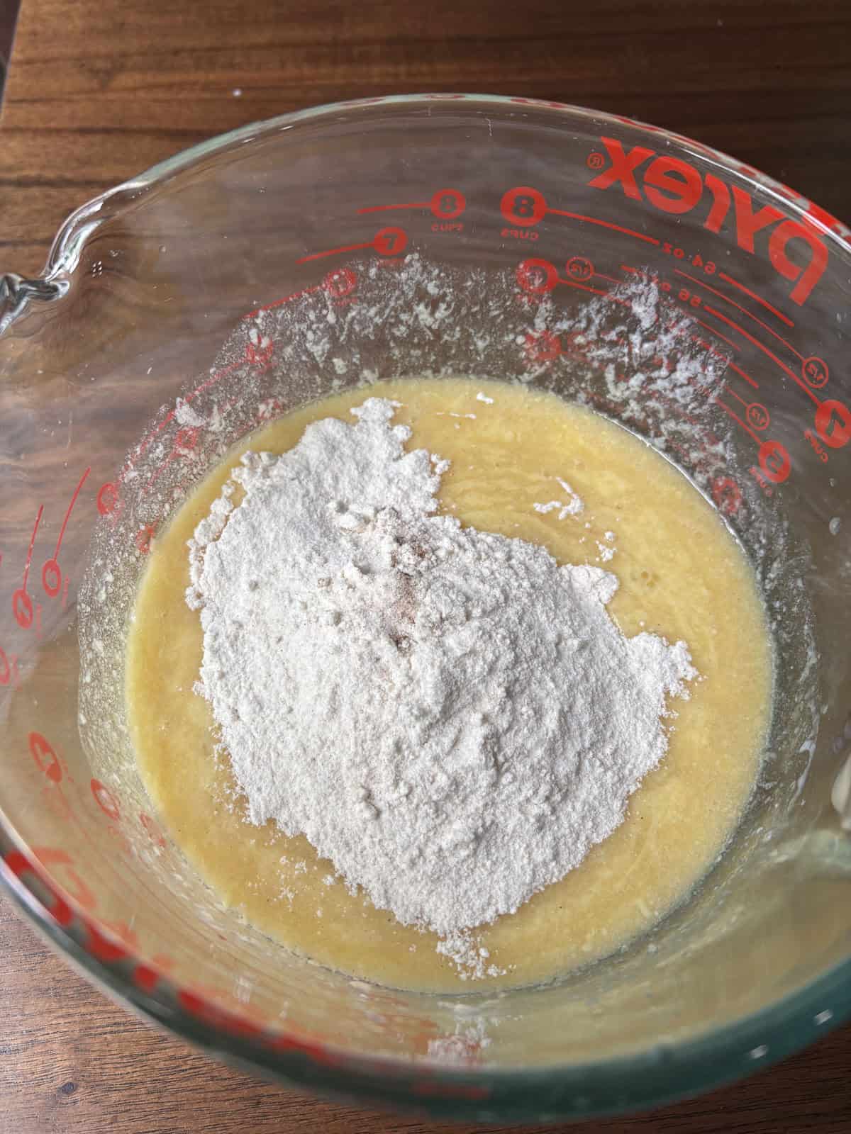 A glass Pyrex measuring cup containing a mixture of beaten eggs and sugar, with a mound of flour on top, sits on a wooden surface, ready to be mixed for baking.
