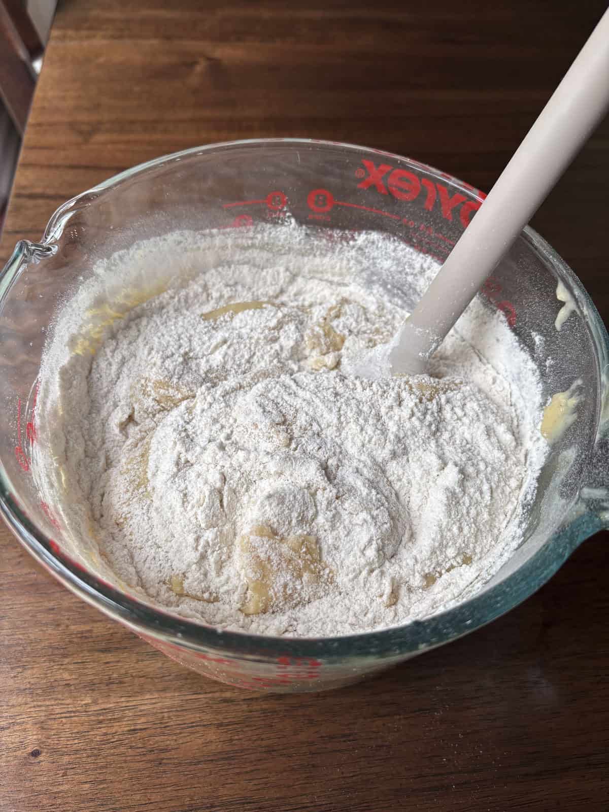 A glass measuring bowl filled with a mixture of flour and wet ingredients is being stirred with a white spatula on a wooden table. The flour is partially mixed into the batter.