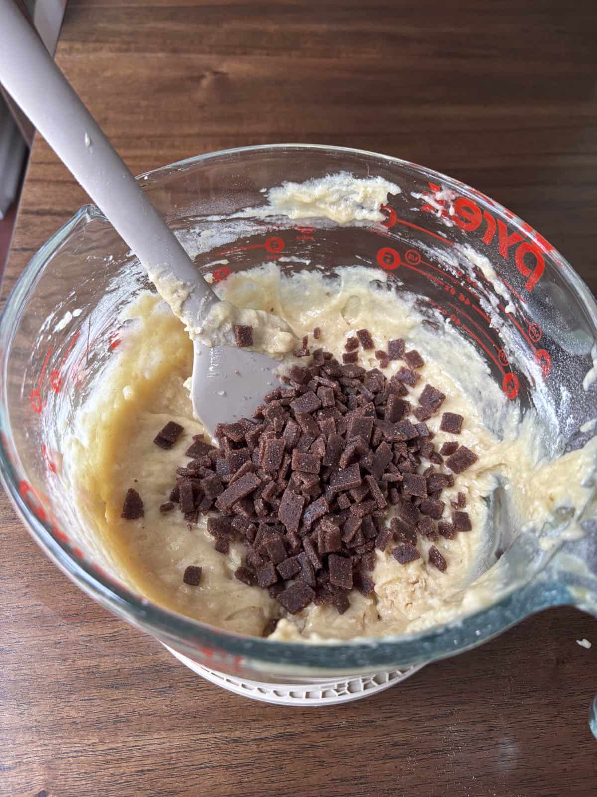 A glass mixing bowl filled with thick batter and topped with a pile of cinnamon chips. A gray spatula rests in the bowl, all set on a wooden surface.