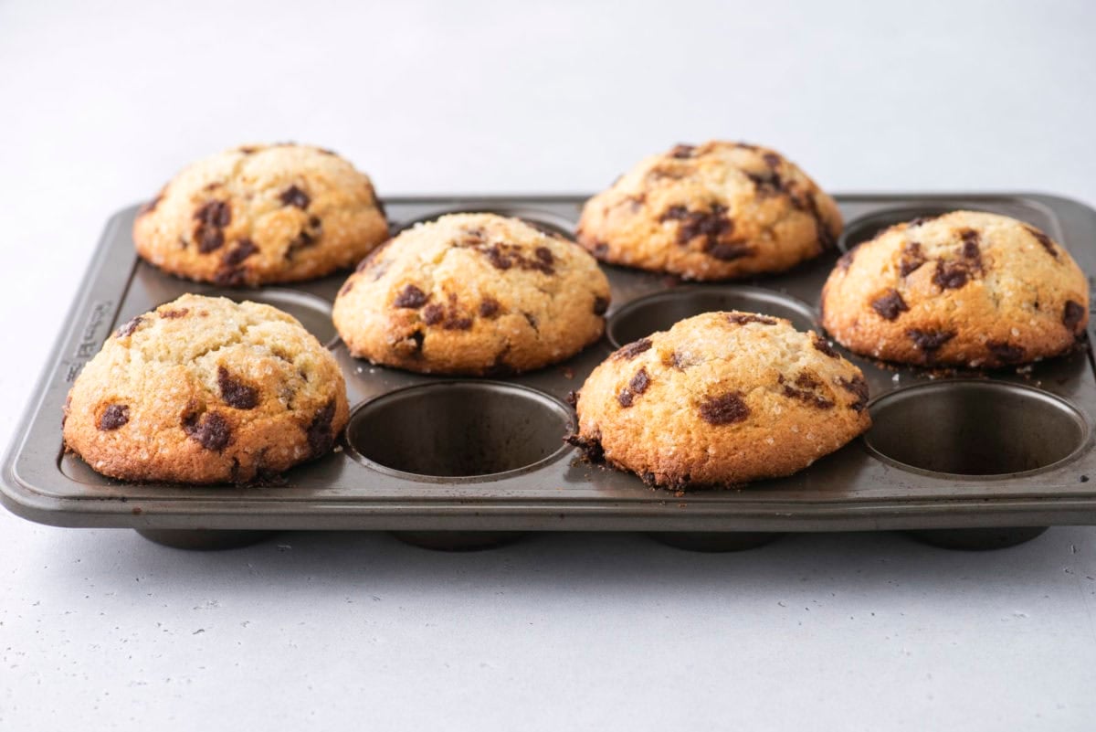 A muffin tray with six golden-brown cinnamon chip muffins, fresh out of the oven, with two empty spaces in the tray, on a light background.