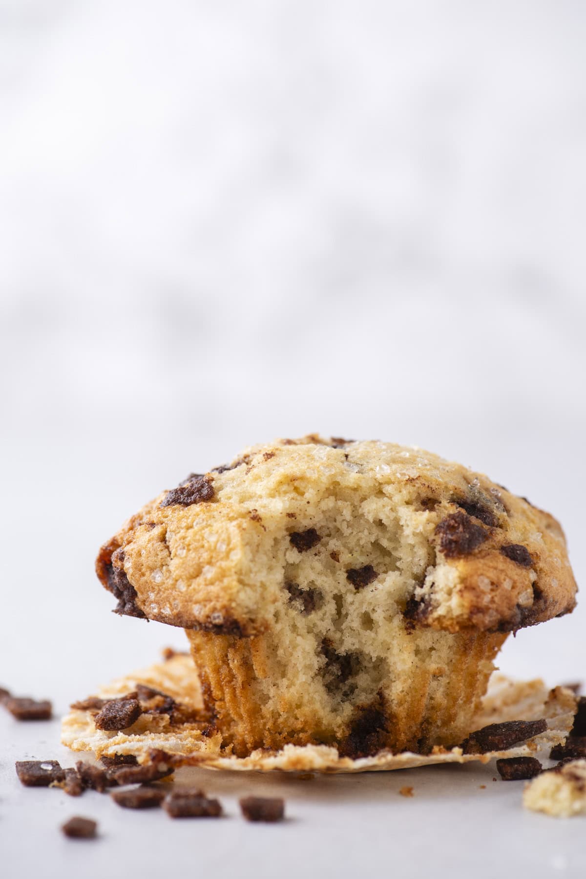 A cinnamon chip muffin with a bite taken out sits on a white surface, partially unwrapped from its paper liner. Crumbs and bits of cinnamon chips are scattered around.