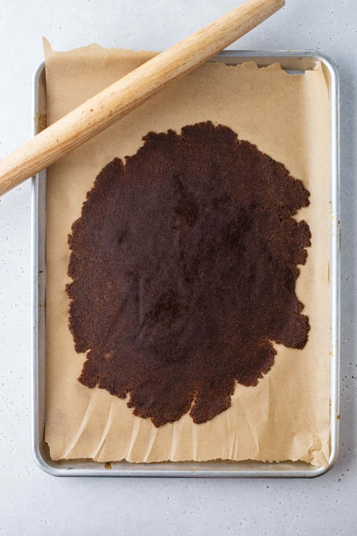 A baking tray lined with parchment paper holds a large, thin sheet of dark dough, partially rolled out. A wooden rolling pin rests diagonally along the top edge of the tray.