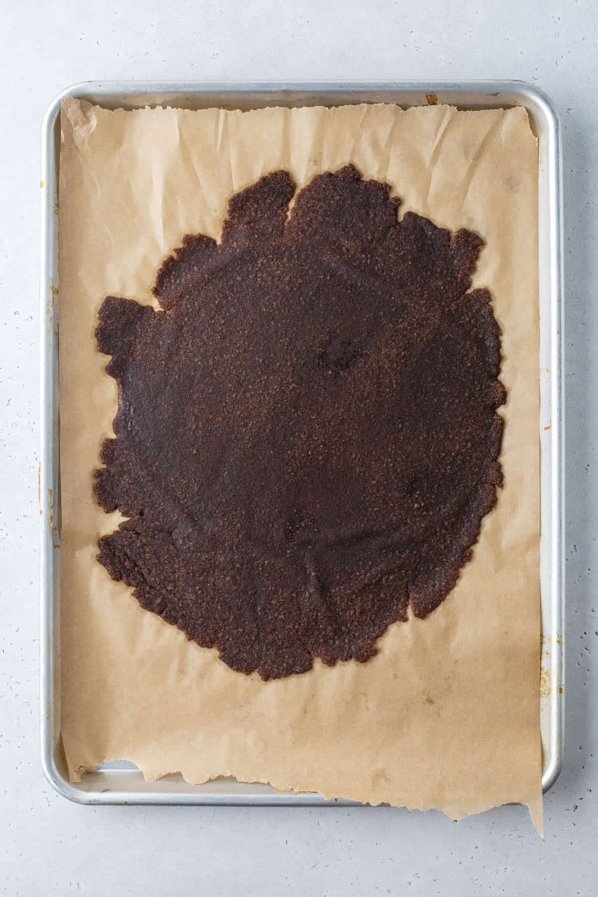 A baking sheet lined with parchment paper holds a large, unevenly spread circle of dark brown dough or mixture. The surface is slightly rough in texture, and the edges are irregular.