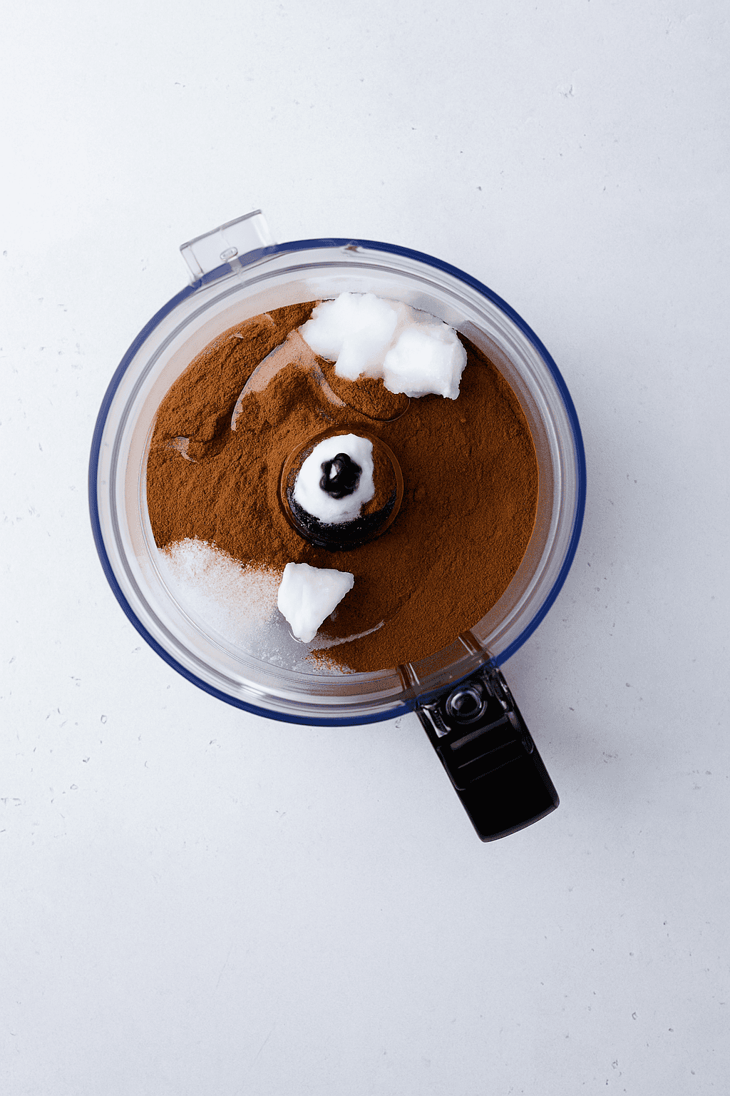 A food processor bowl containing ground cinnamon, granulated sugar, and several dollops of solid coconut oil on a light gray surface.