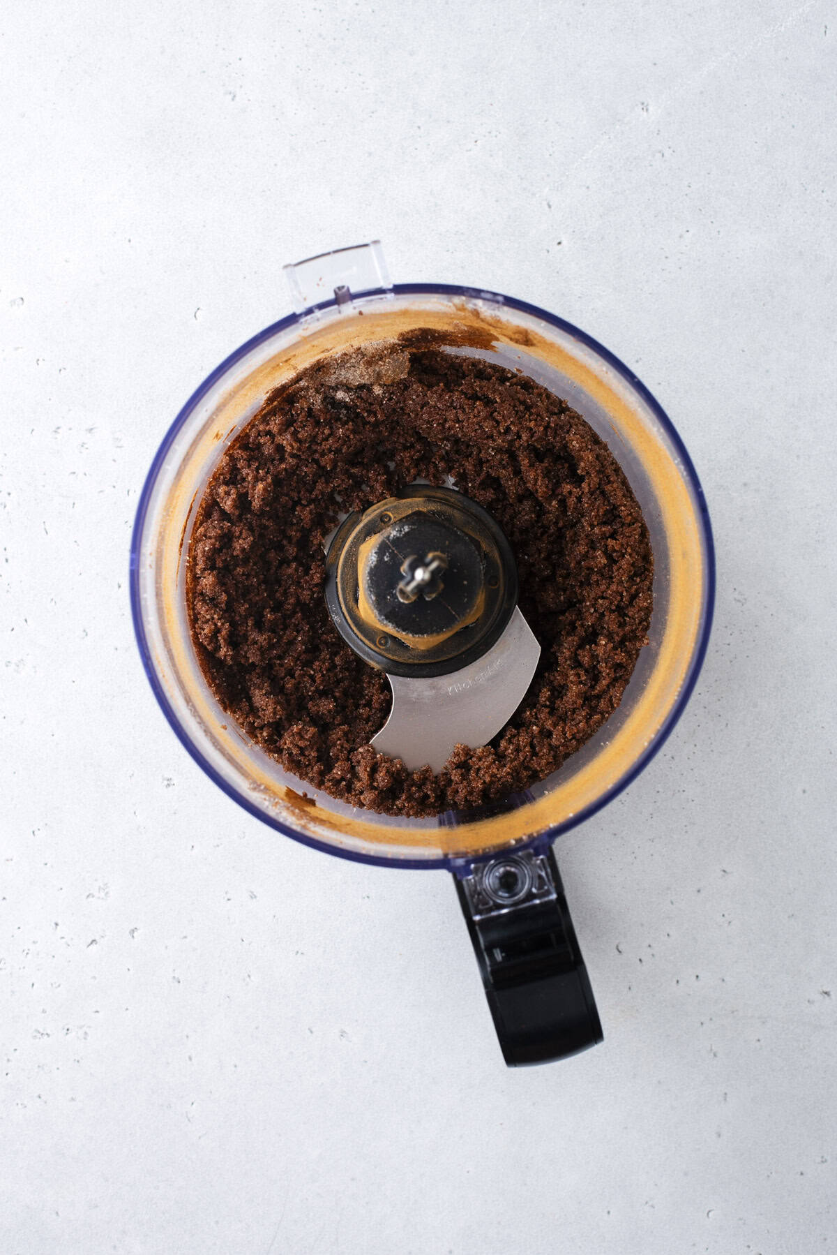 A food processor bowl filled with blended brown mixture, viewed from above on a light gray surface.