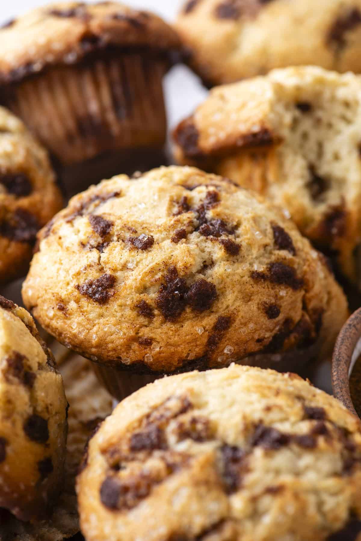 Close-up of cinnamon chip muffins with golden, slightly crisp tops and visible cinnamon pieces, one muffin partially broken open to show the soft, moist inside.