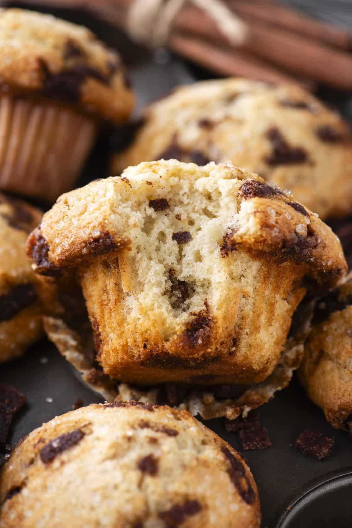 A close-up of cinnamon chip muffins, with one muffin cut in half to show its moist, fluffy texture and cinnamon chunks inside. Other whole muffins and cinnamon sticks are in the background.
