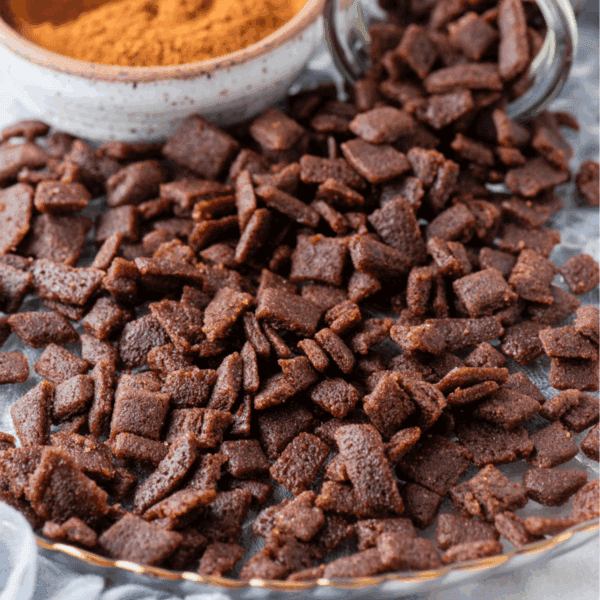 A close-up of homemade cinnamon chips spread on a plate, with a bowl of cinnamon powder in the background. The cereal appears crunchy and is coated with a brown, sugary cinnamon mixture.