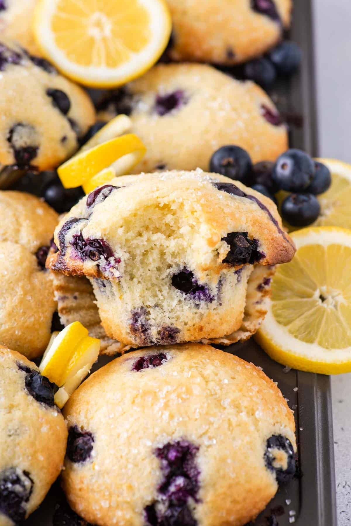 A batch of blueberry muffins on a baking tray, with one muffin in the center missing a bite. Fresh blueberries and lemon slices are scattered around the muffins, highlighting the ingredients.