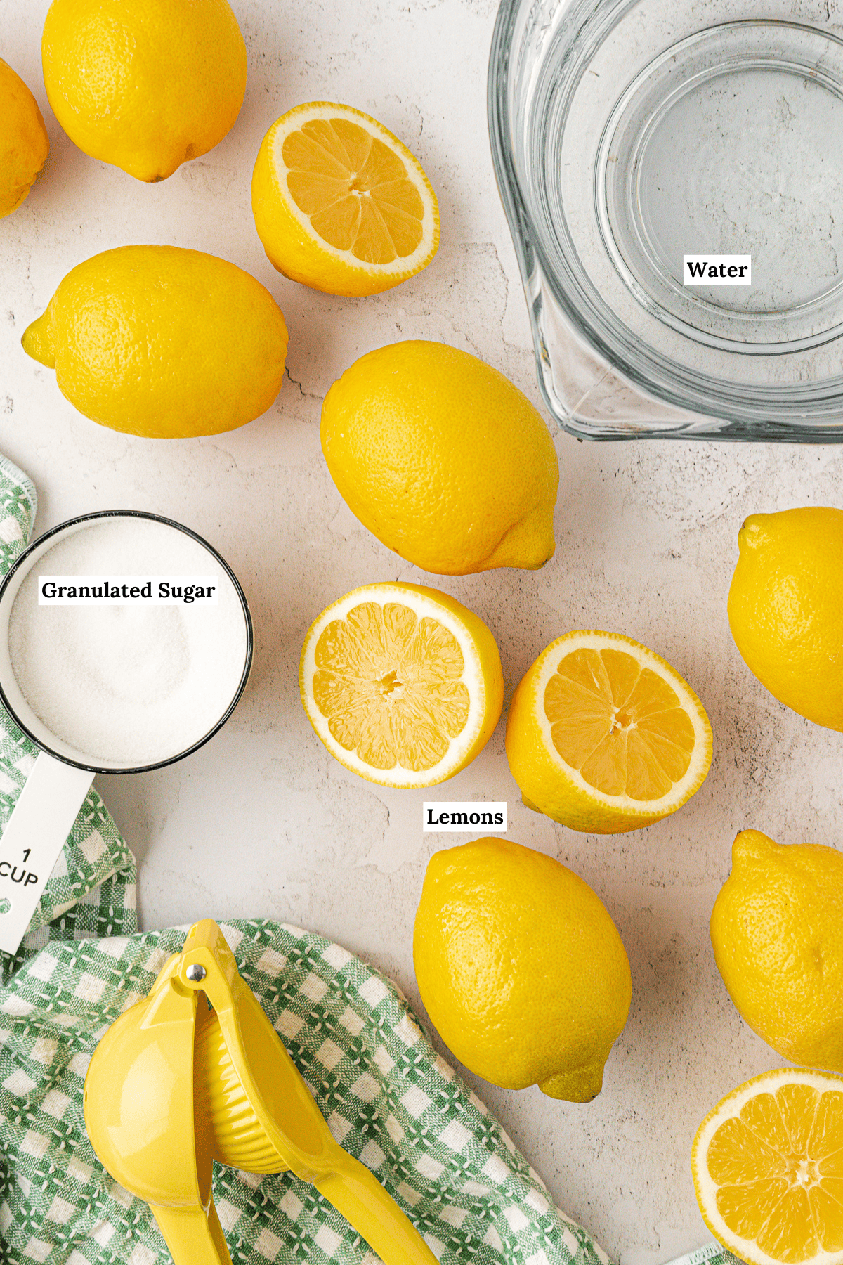 Overhead view of whole and halved lemons, a bowl of water, a measuring cup of granulated sugar, a yellow citrus juicer, and a green checkered cloth on a white surface. Some items are labeled.