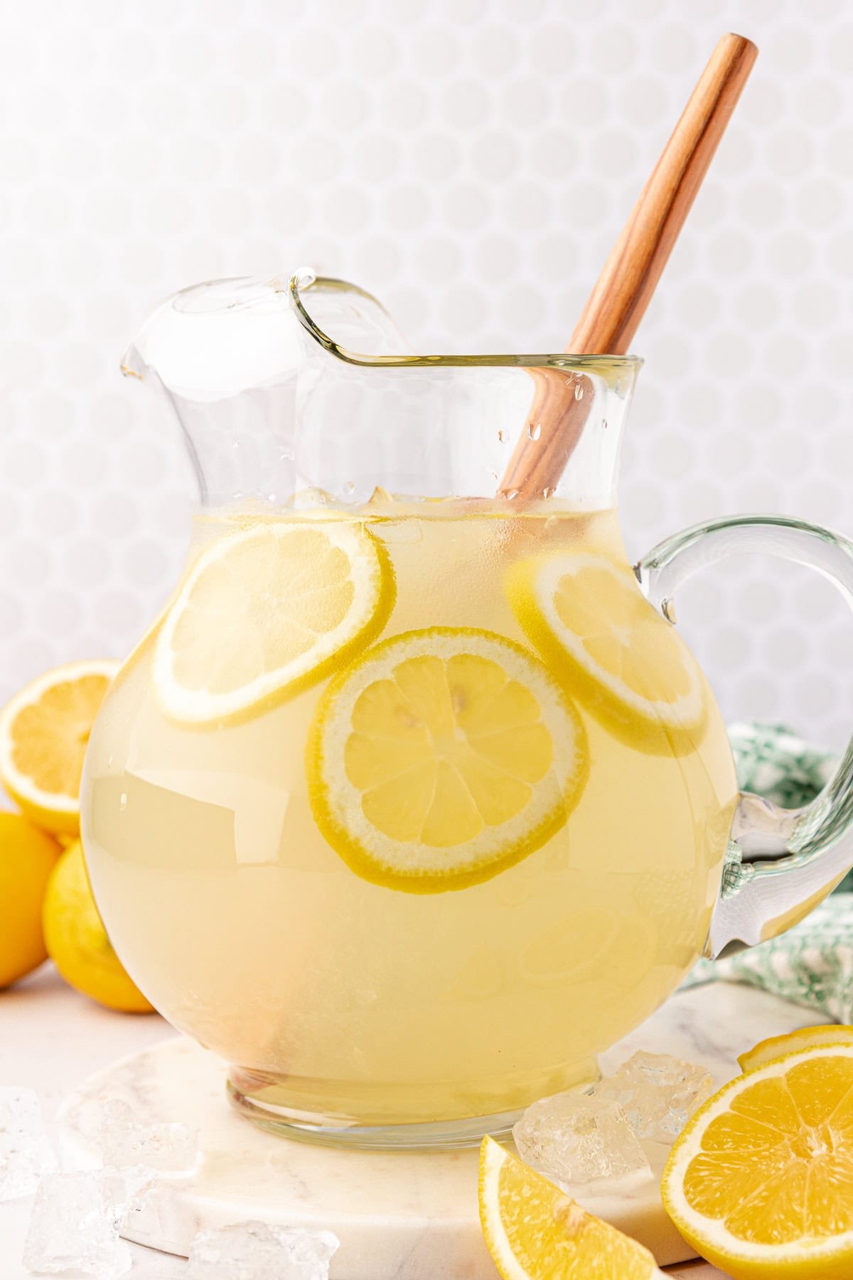 A glass pitcher filled with lemonade and lemon slices, with a wooden spoon inside. Ice cubes and lemon wedges are scattered around the pitcher on a white surface.