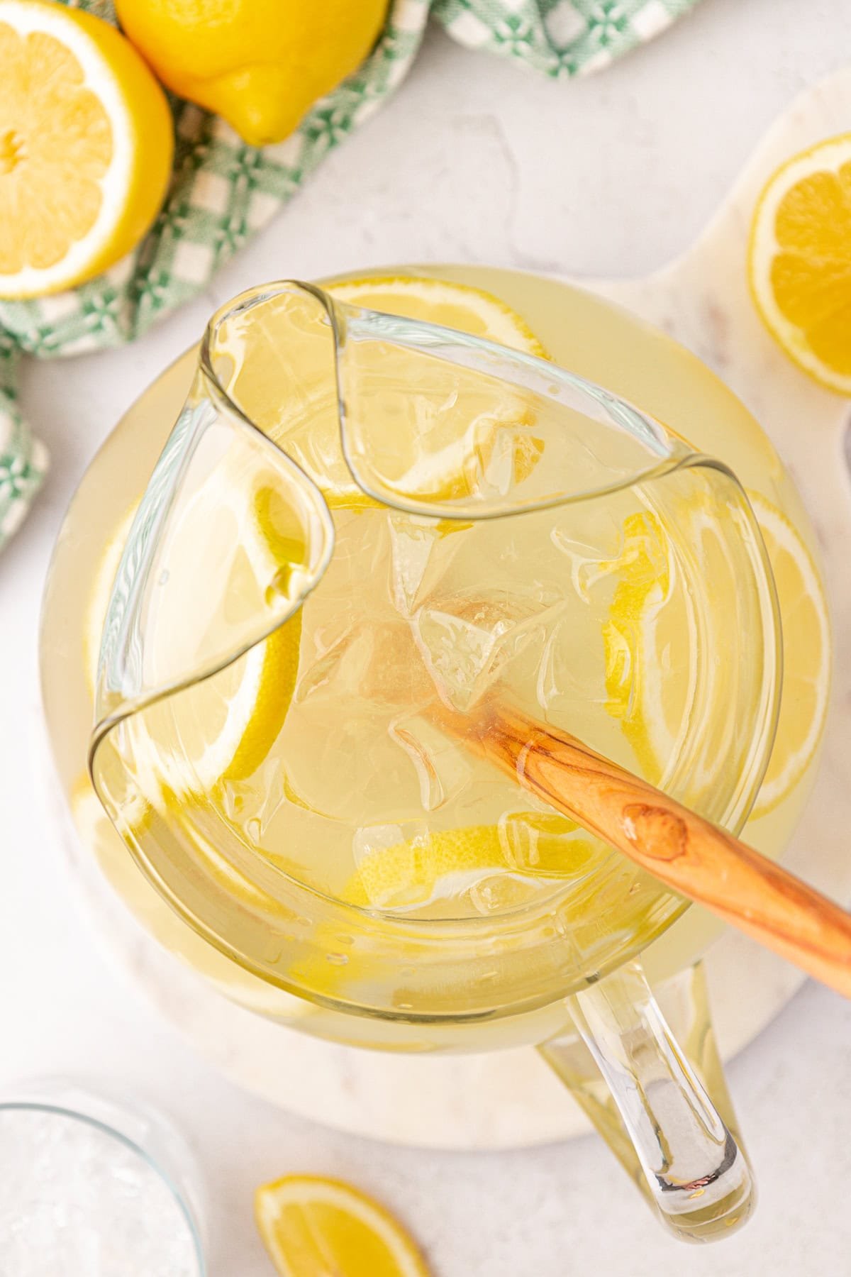 A top view of a glass pitcher filled with iced lemonade and lemon slices, being stirred with a wooden spoon. Fresh lemon halves and a green checkered cloth are visible in the background.