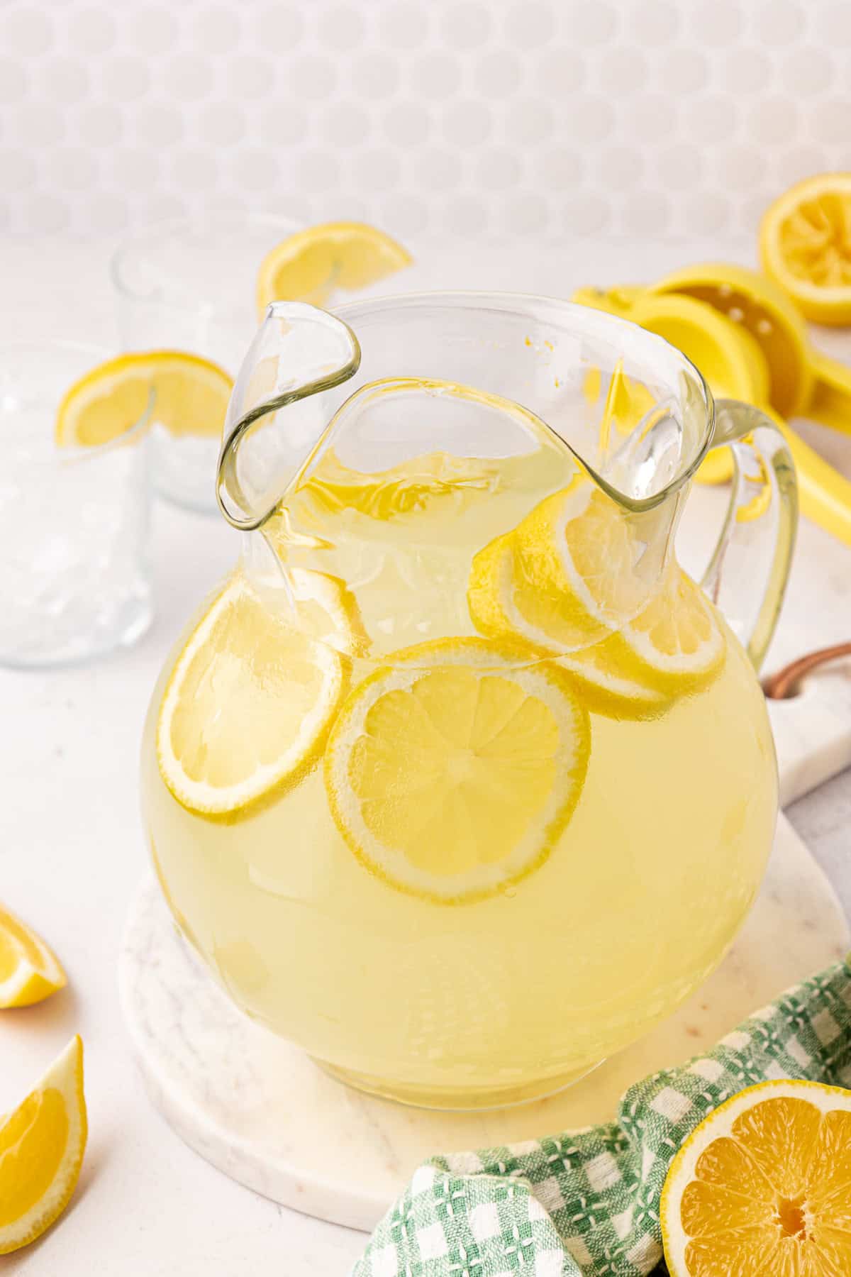 A glass pitcher of lemonade with floating lemon slices sits on a marble surface. Lemon wedges, a green checkered towel, and a yellow citrus squeezer are nearby.