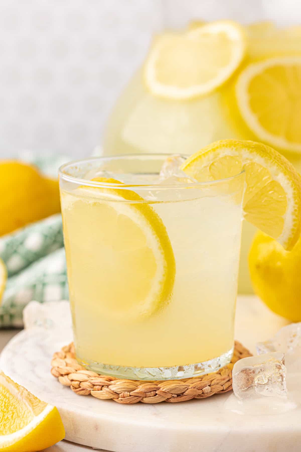 A glass of lemonade with ice and a lemon slice garnish sits on a woven coaster, with a pitcher of lemonade and additional lemon slices in the background.