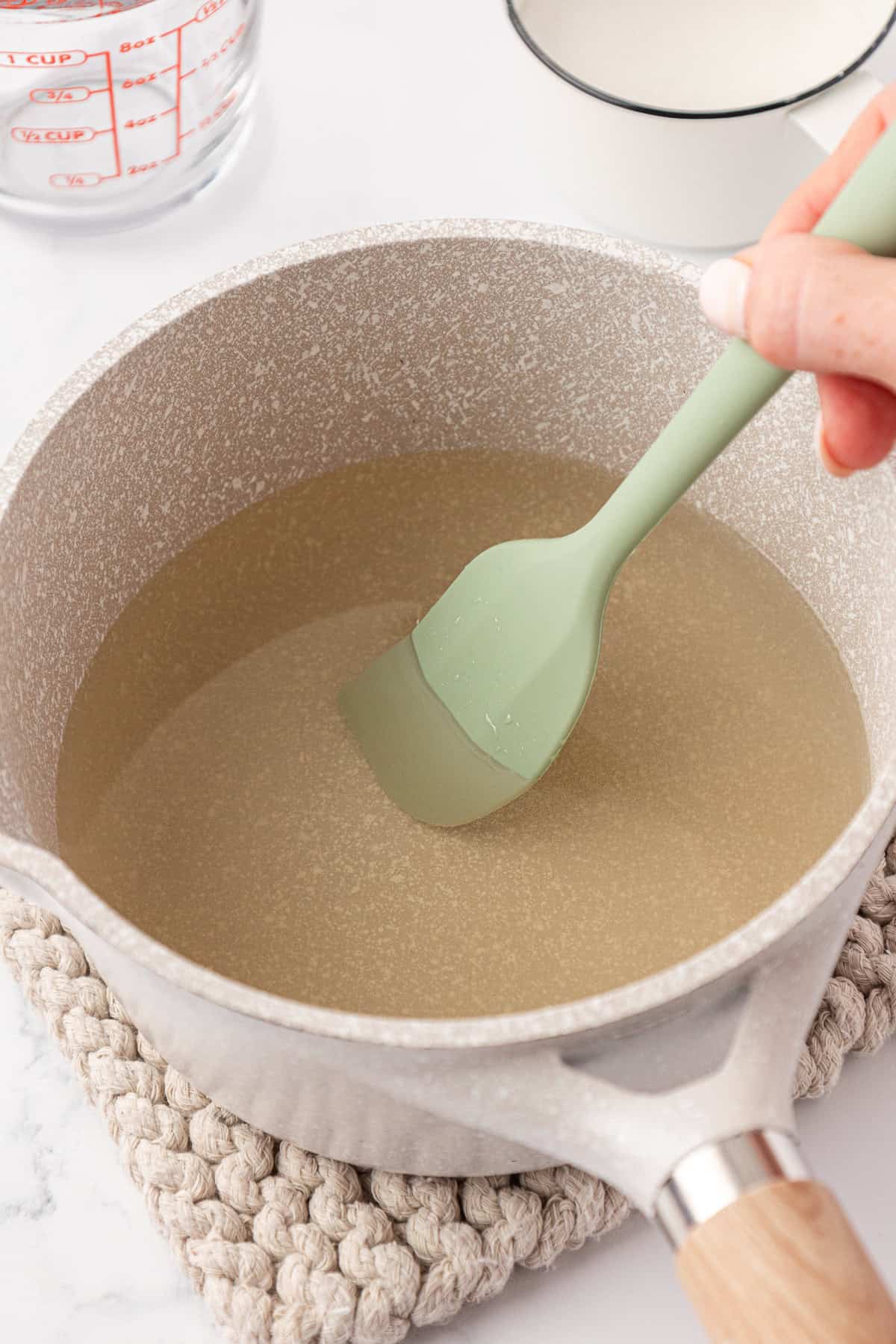 A hand stirs a clear liquid in a saucepan with a green spatula. The saucepan sits on a textured mat, and a glass measuring cup is visible nearby.