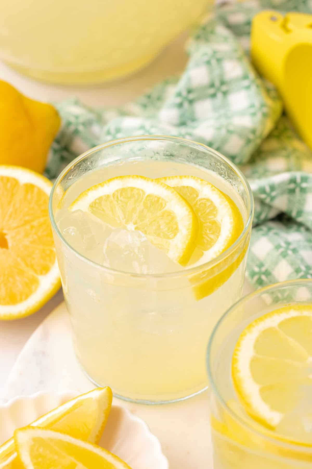 A glass of lemonade with ice and lemon slices, surrounded by more lemon wedges and a green checkered cloth in the background.