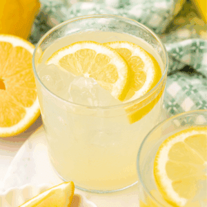 A glass of lemonade with ice and lemon slices, placed on a white surface next to a halved lemon and a patterned green and white cloth in the background.