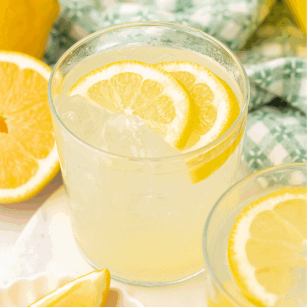 A glass of lemonade with ice and lemon slices, placed on a white surface next to a halved lemon and a patterned green and white cloth in the background.