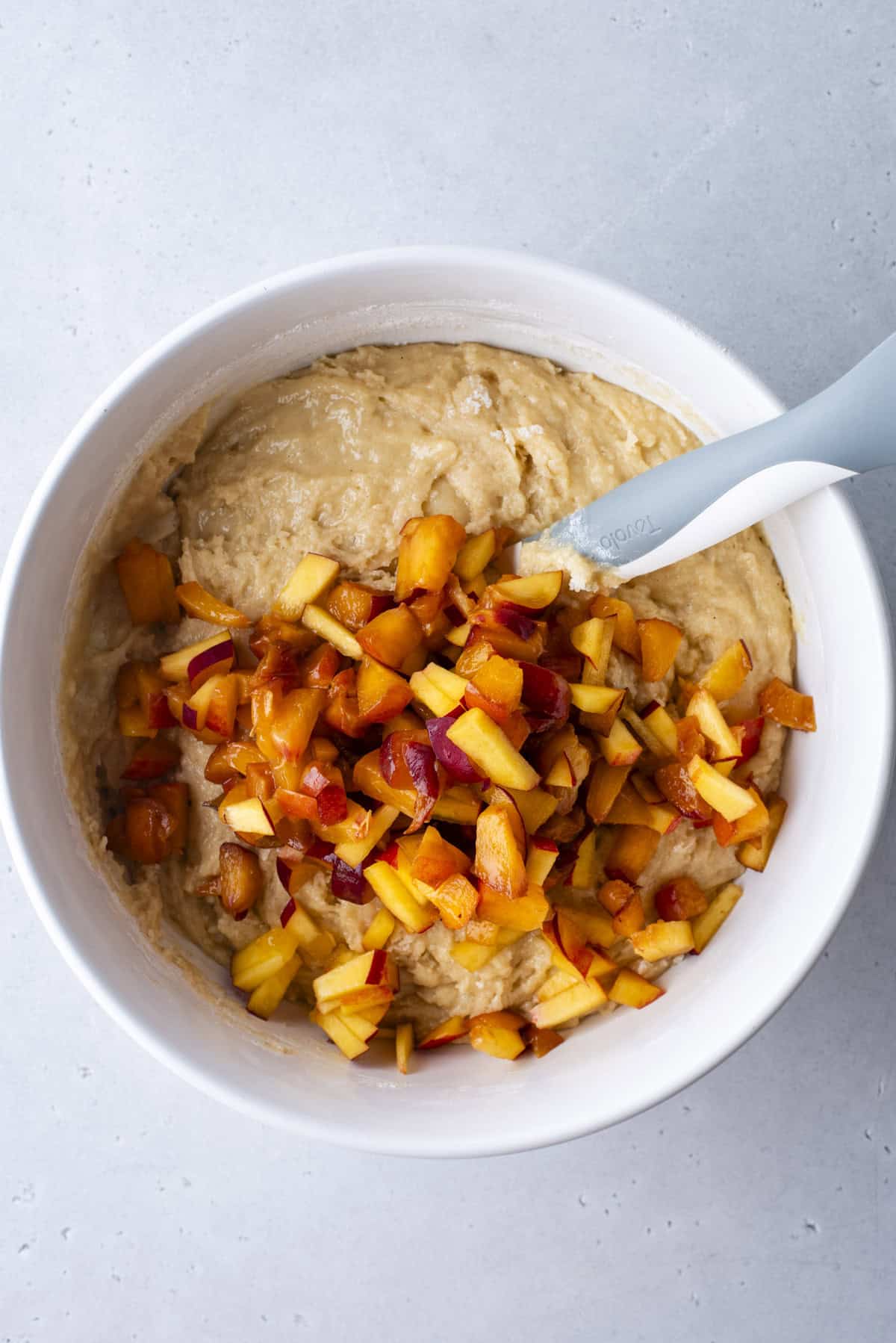 A white mixing bowl with banana bread batter, topped with chopped peaches and a sprinkle of cinnamon, with a gray spatula resting inside.