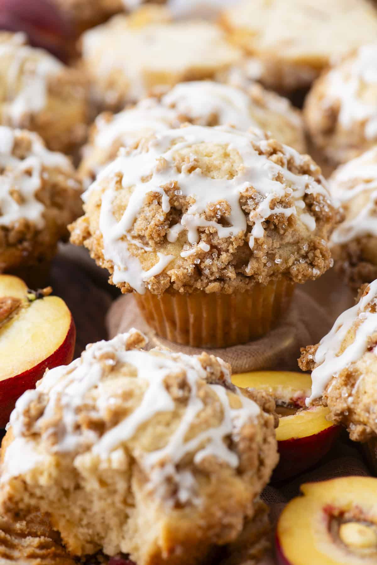 A close-up of peach muffins topped with crumbly streusel and white icing, surrounded by fresh peach slices. The muffins are golden brown with a generous drizzle of icing.