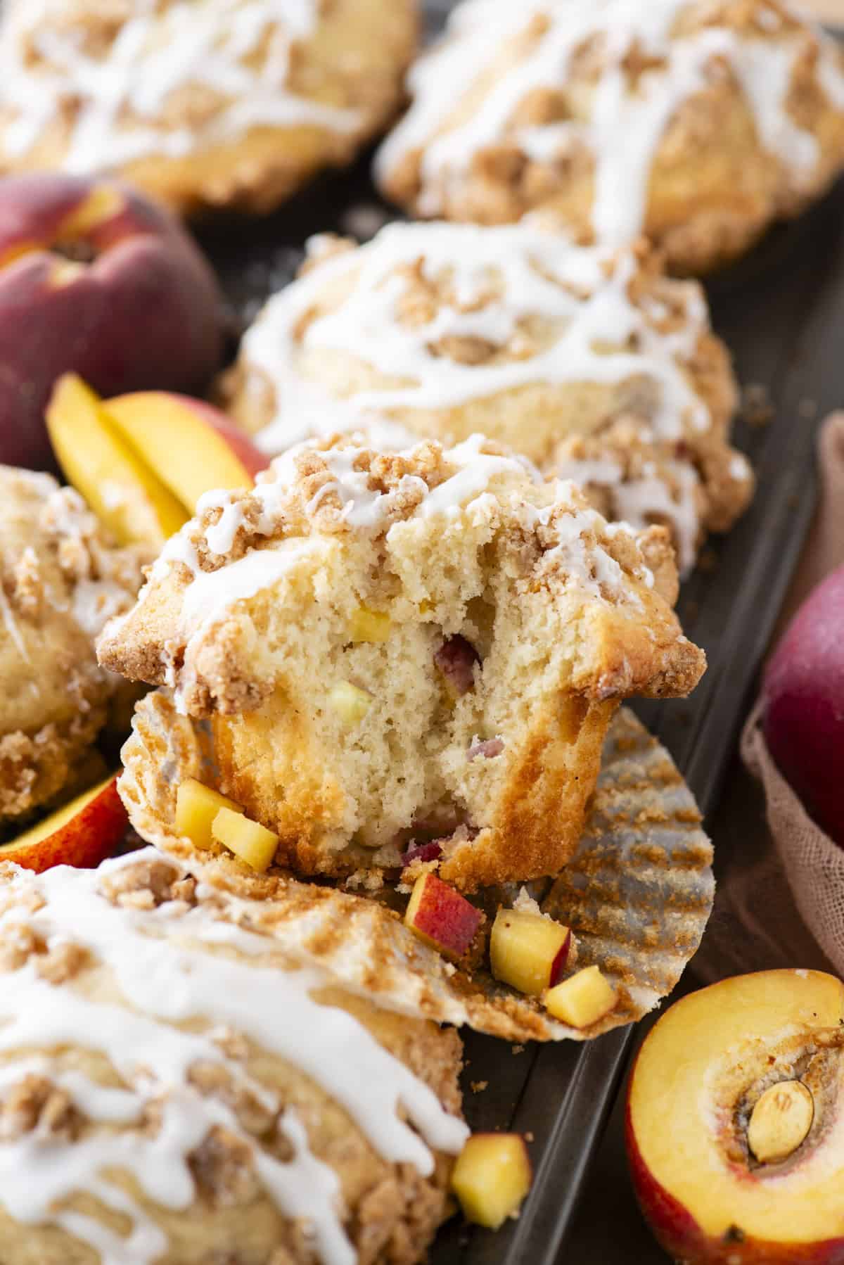 A close-up of peach muffins with crumb topping and white icing drizzle, one muffin cut open to reveal chunks of fresh peach inside. Sliced peaches and whole peaches are placed around the muffins.