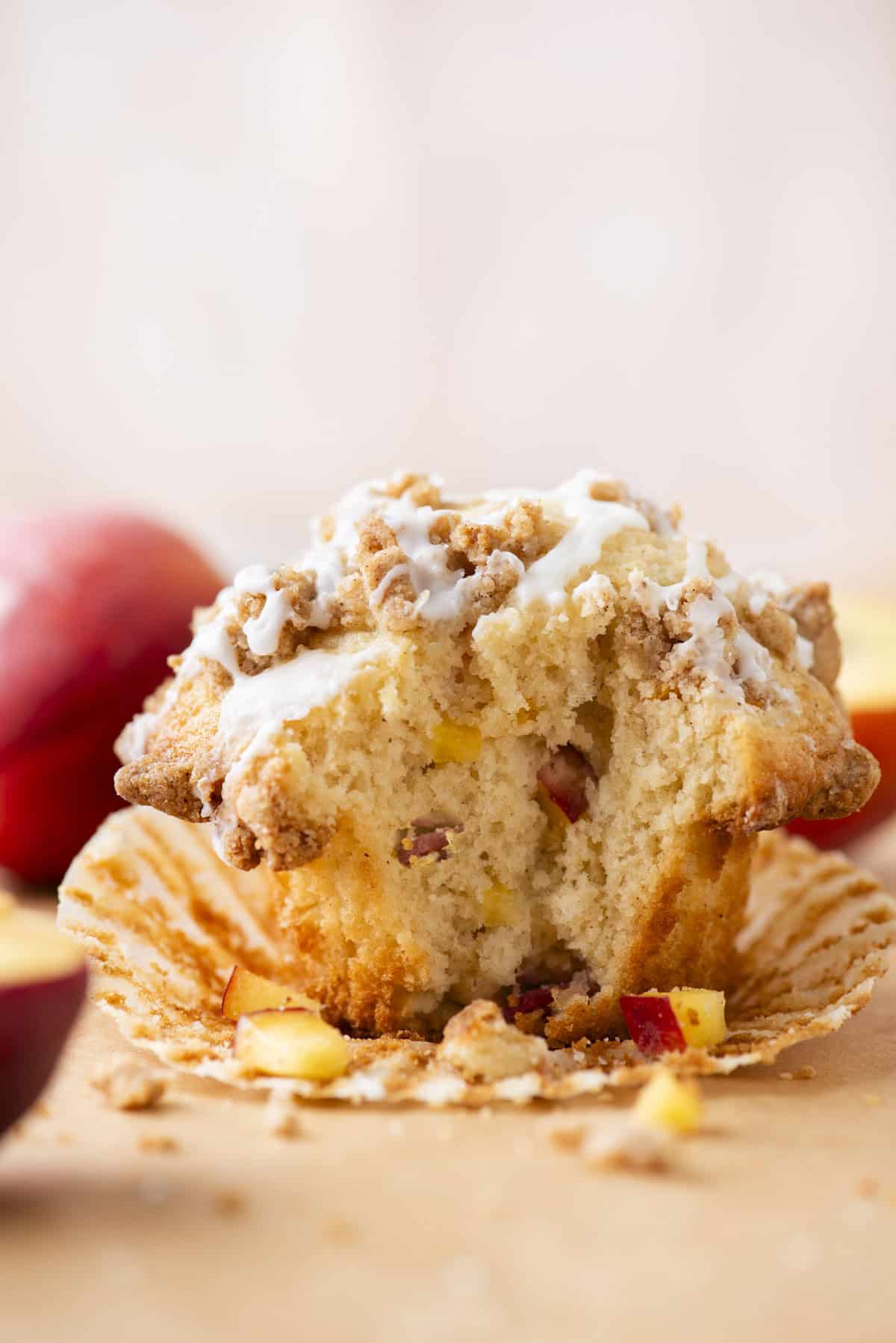 A close-up of a peach muffin with a crumb topping and white icing, partially unwrapped in a muffin liner. Crumbs and small pieces of peach are scattered around the muffin.