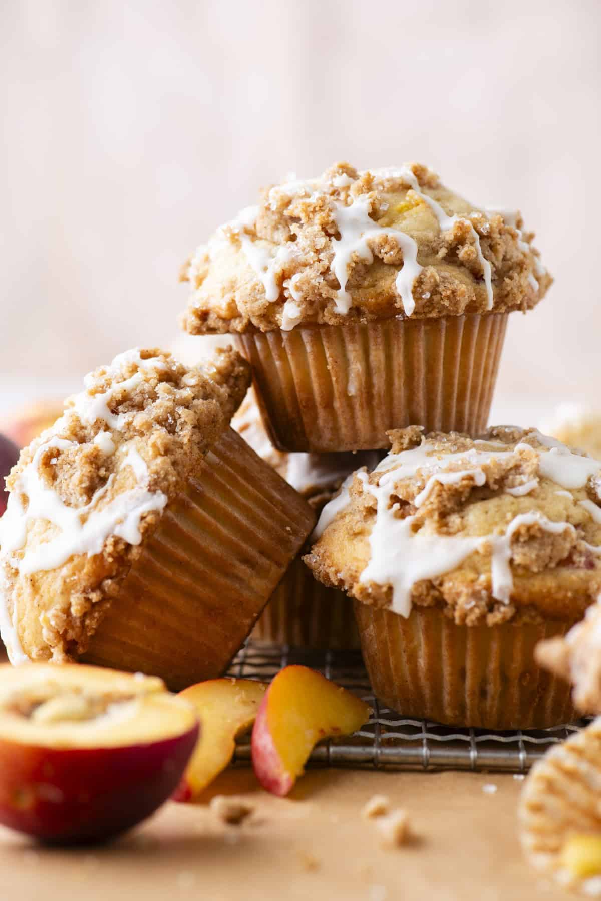 Three peach muffins with crumb topping and white icing are stacked on a cooling rack. In the foreground, there are peach slices, and the background is softly blurred.