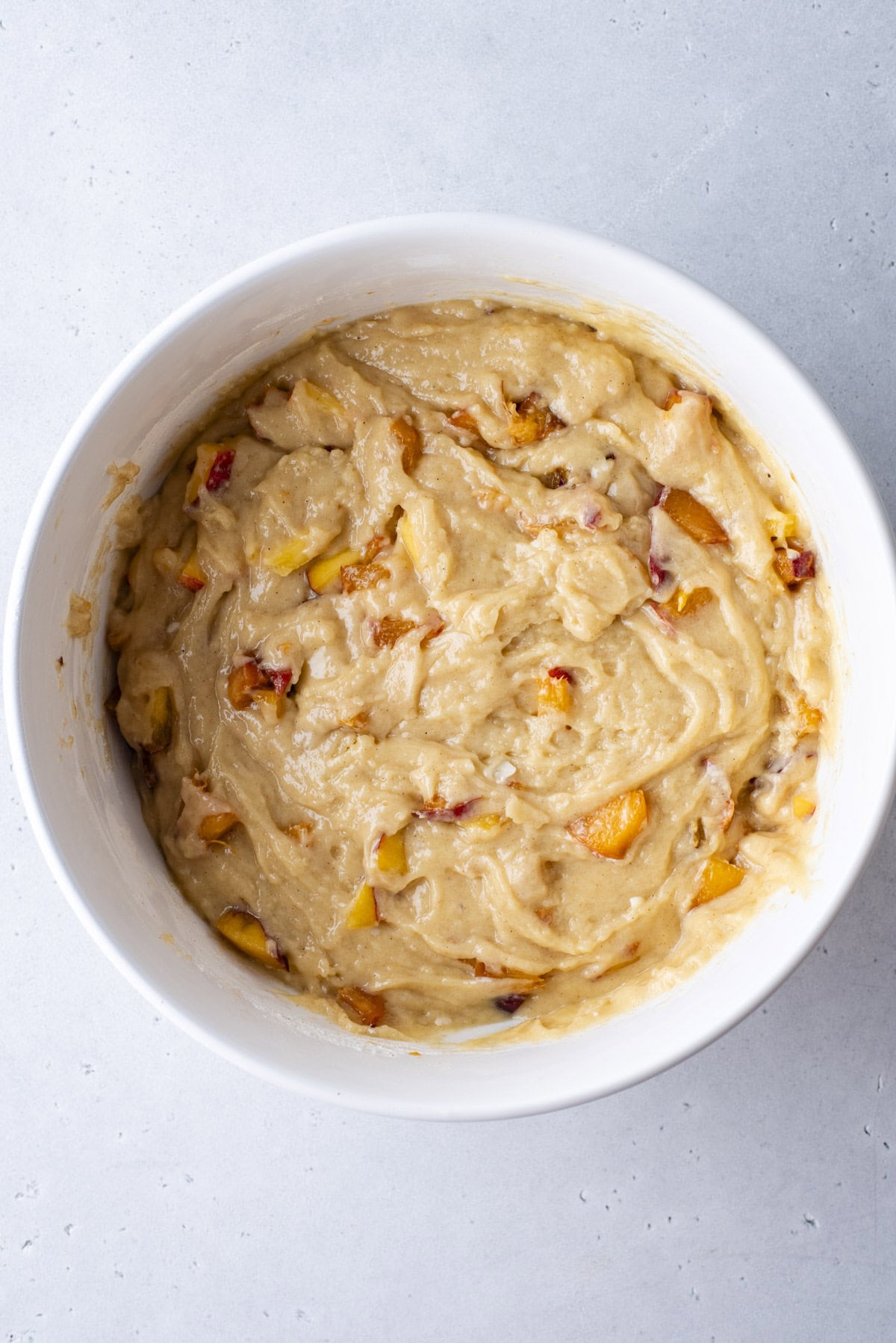 A white bowl filled with thick, creamy cake batter mixed with visible chunks of peaches, sitting on a light gray countertop.