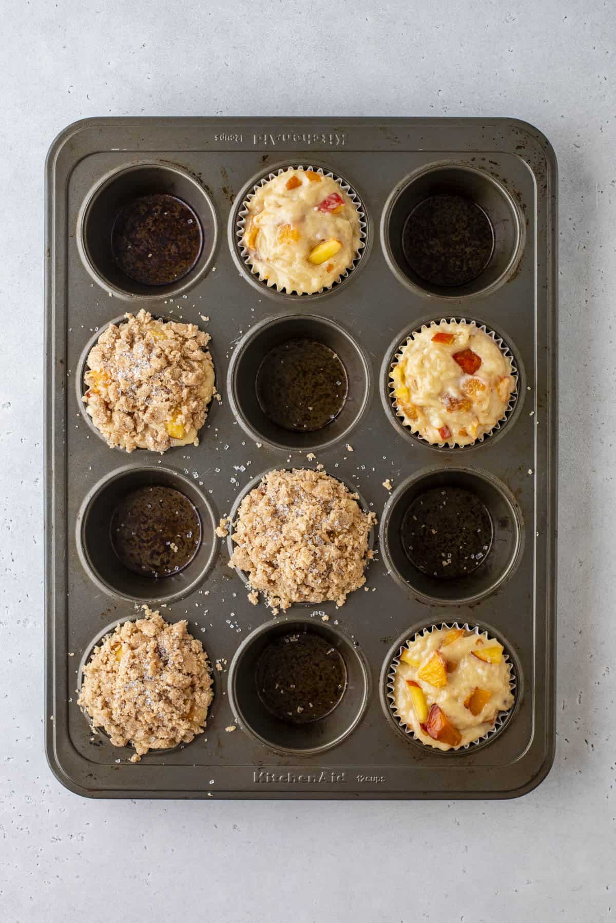 A muffin tin holds six paper liners filled with unbaked muffin batter, some topped with crumbly streusel and others with visible fruit pieces. The other six cavities are empty and visibly greased.
