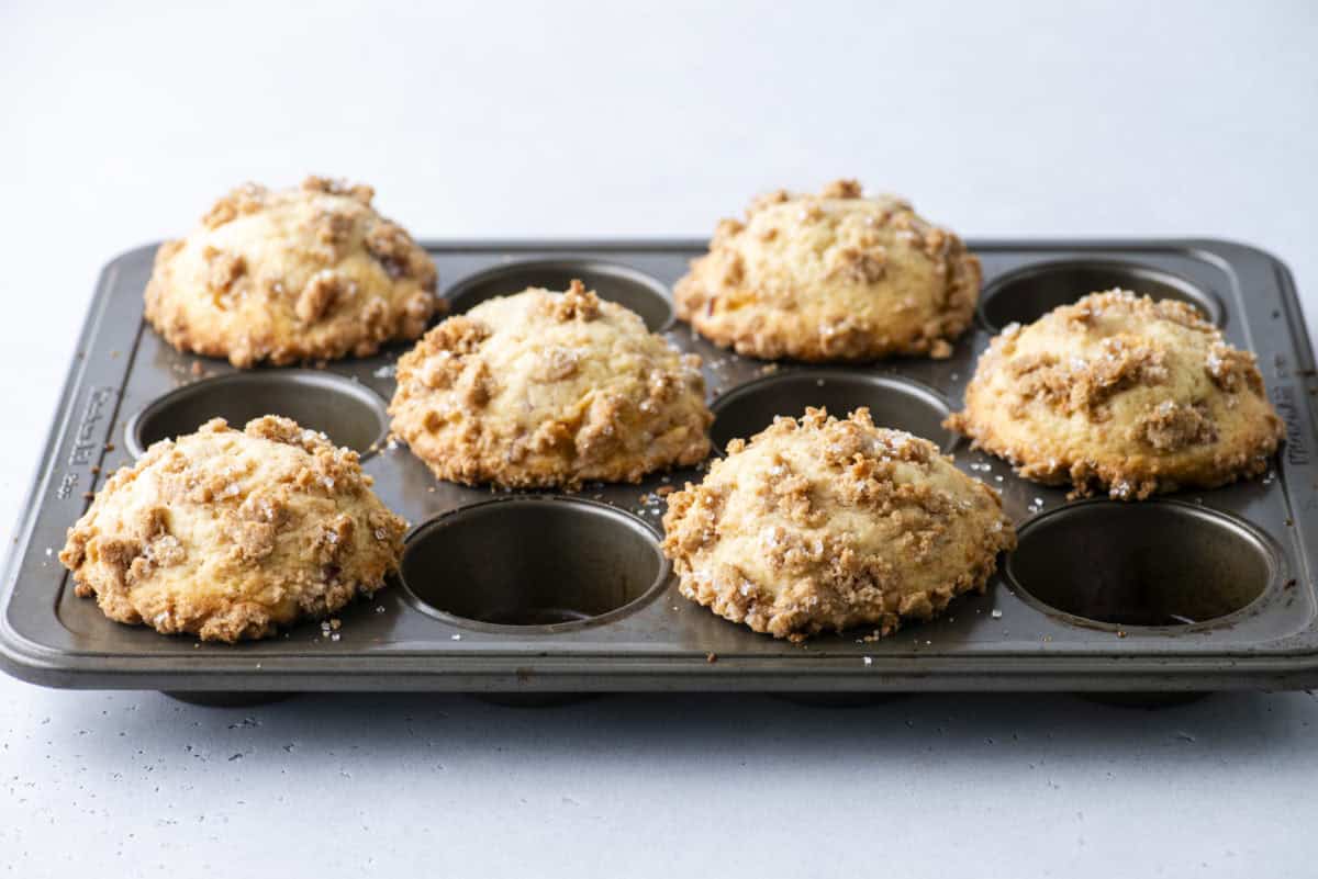 A muffin tin holding six golden-brown, crumbly-topped muffins with two empty spaces, set on a light gray surface.