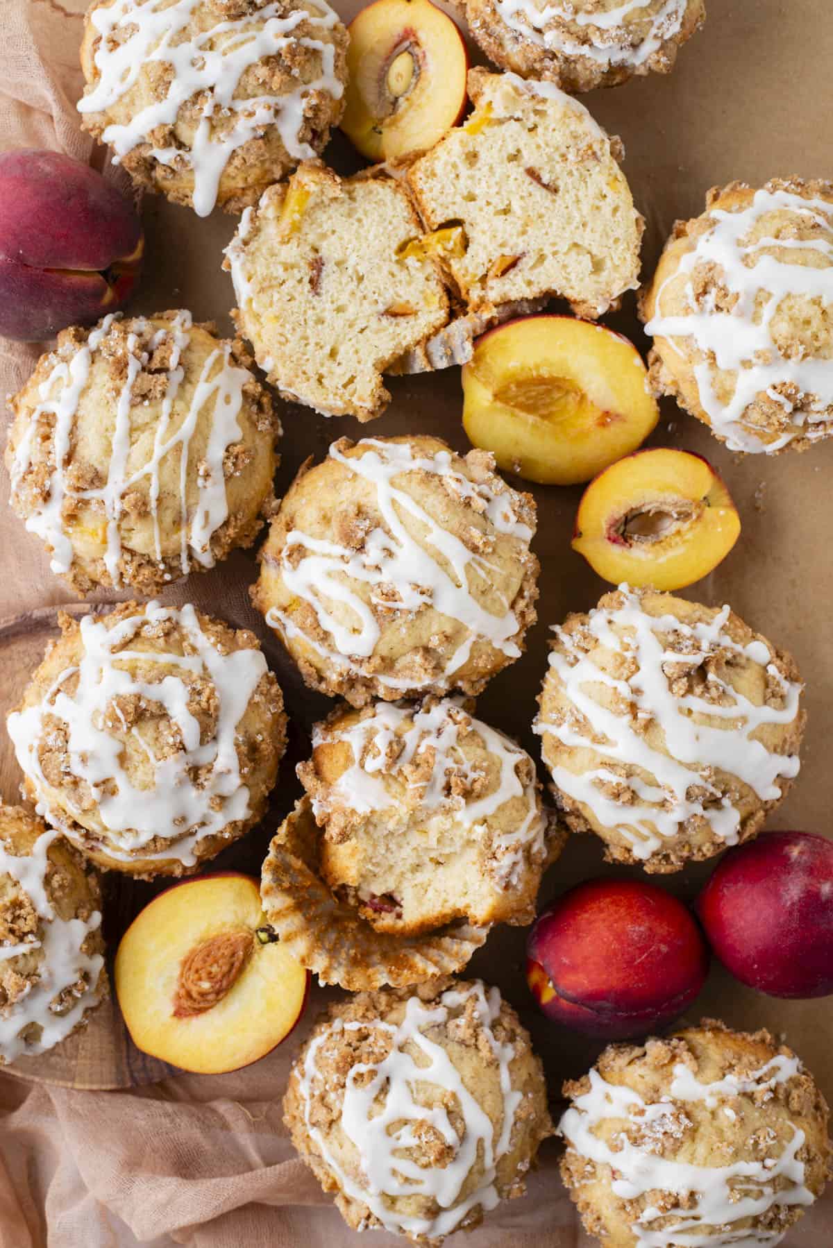 A top view of streusel-topped muffins drizzled with white icing, surrounded by split and whole peaches and plums on a brown parchment surface. One muffin is cut open to reveal a soft, moist interior with fruit pieces.