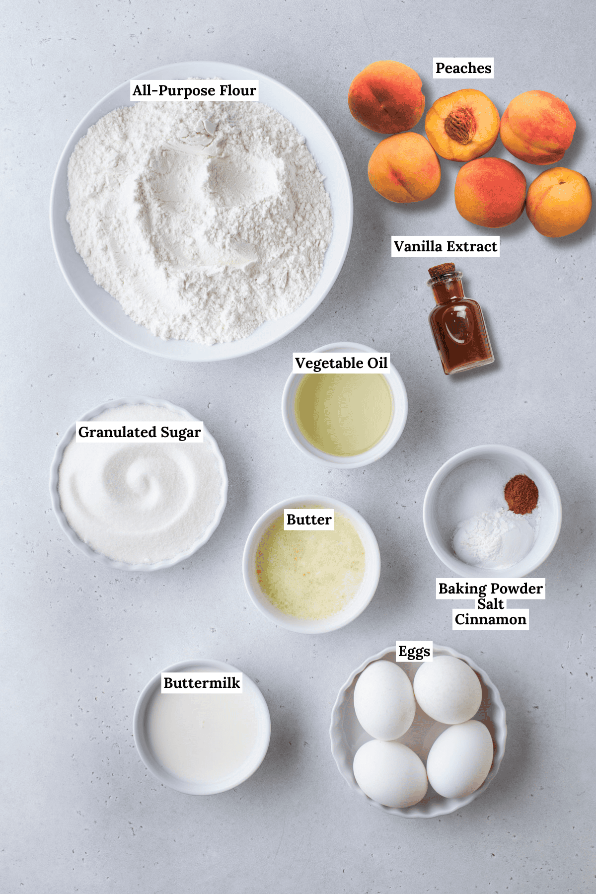 Bowls containing all-purpose flour, granulated sugar, buttermilk, eggs, vegetable oil, melted butter, baking powder, salt, cinnamon, a bottle of vanilla extract, and whole peaches are arranged on a light surface.