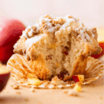 A close-up of a partially eaten muffin with visible apple chunks, drizzled with white icing and topped with crumbly streusel, sitting on a peeled muffin wrapper with peach slices in the background.
