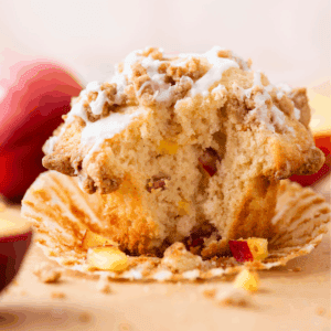 A close-up of a partially eaten muffin with visible apple chunks, drizzled with white icing and topped with crumbly streusel, sitting on a peeled muffin wrapper with peach slices in the background.