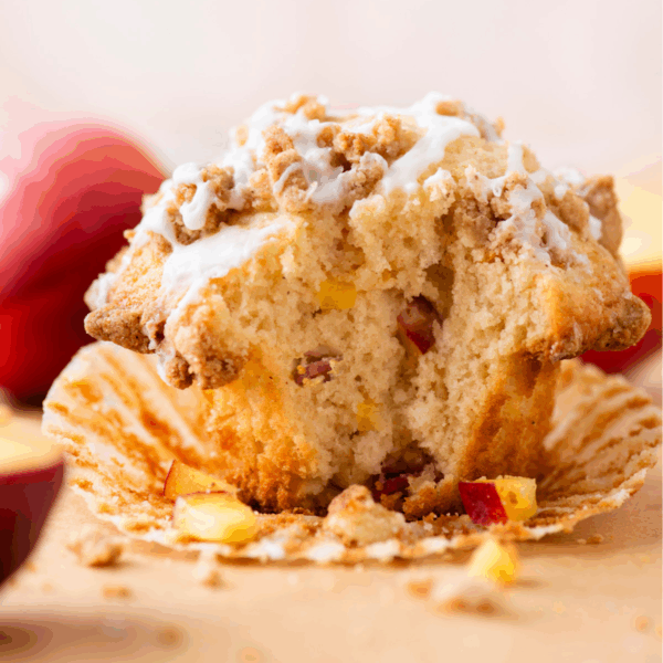 A close-up of a partially eaten muffin with visible apple chunks, drizzled with white icing and topped with crumbly streusel, sitting on a peeled muffin wrapper with peach slices in the background.