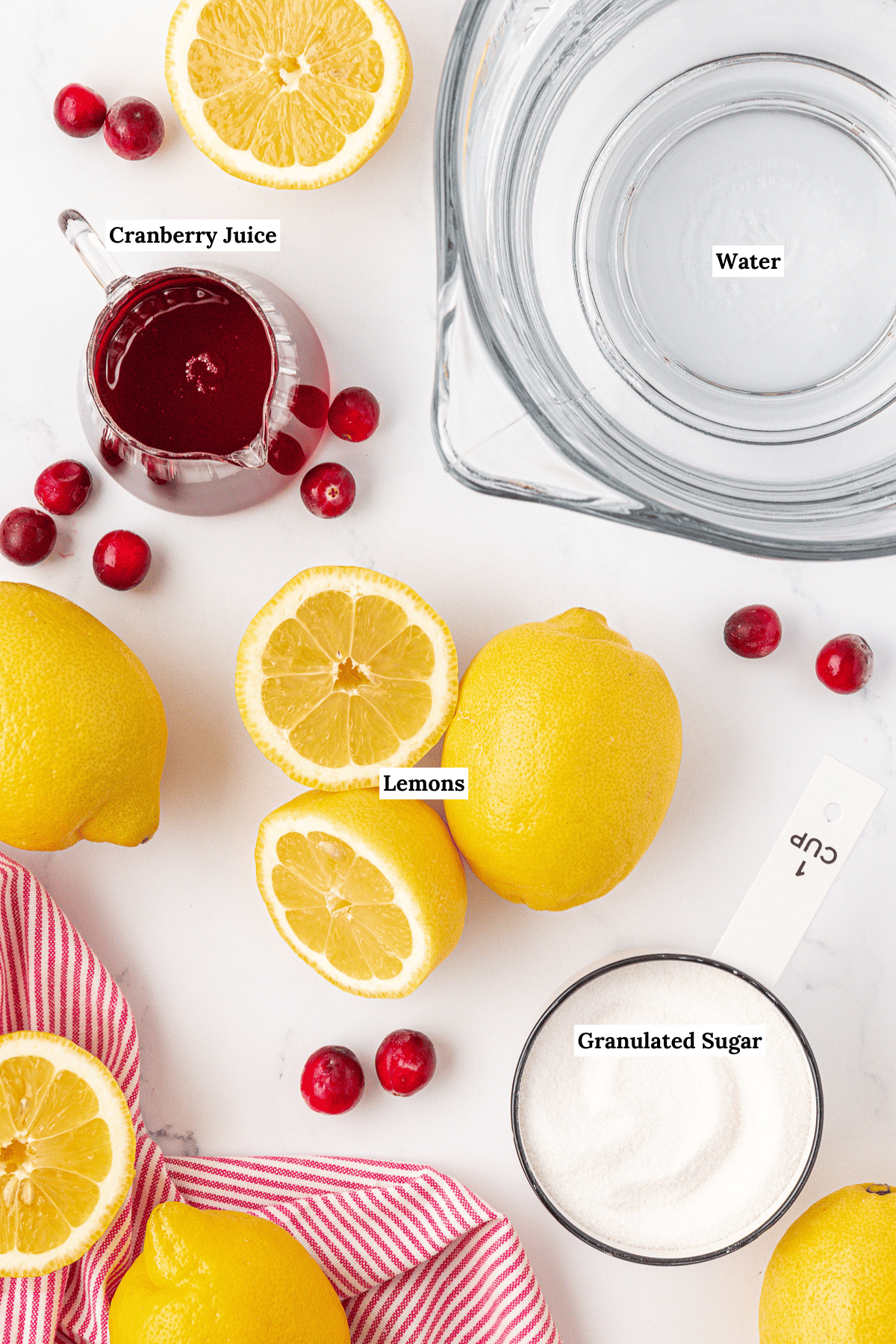 Top-down view of ingredients for cranberry lemonade: a glass of water, cranberry juice, whole and halved lemons, granulated sugar in a measuring cup, loose cranberries, and a red-striped cloth on a white surface. Labels identify each item.
