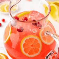 A glass pitcher filled with pink lemonade, ice cubes, lemon slices, and cranberries sits on a white surface. Lemon slices and cranberries are scattered around. Text at the top reads Pink Lemonade.