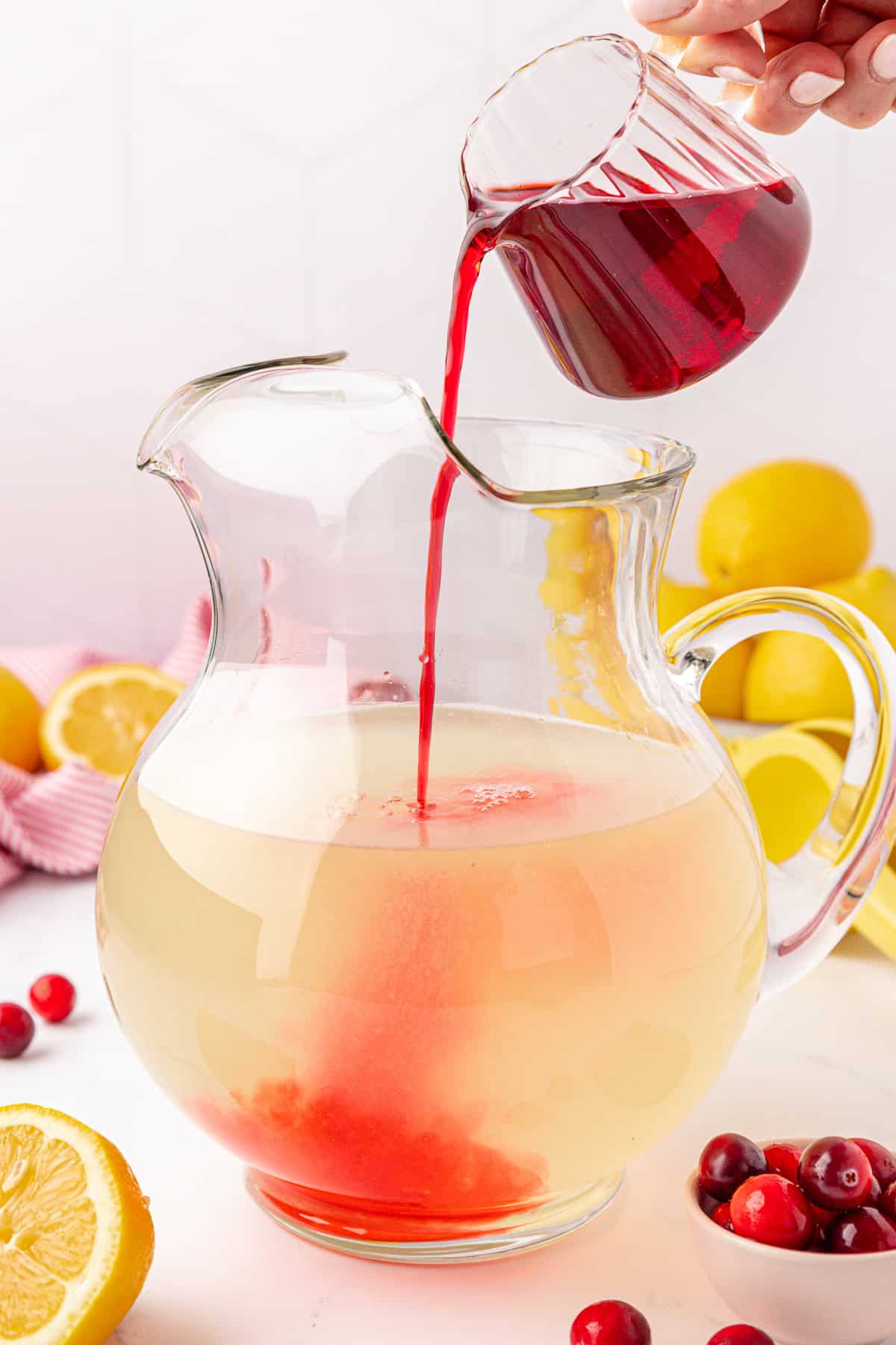 A hand pours red liquid from a small glass pitcher into a large clear pitcher of lemonade. Lemons and cranberries are scattered around on a white surface.