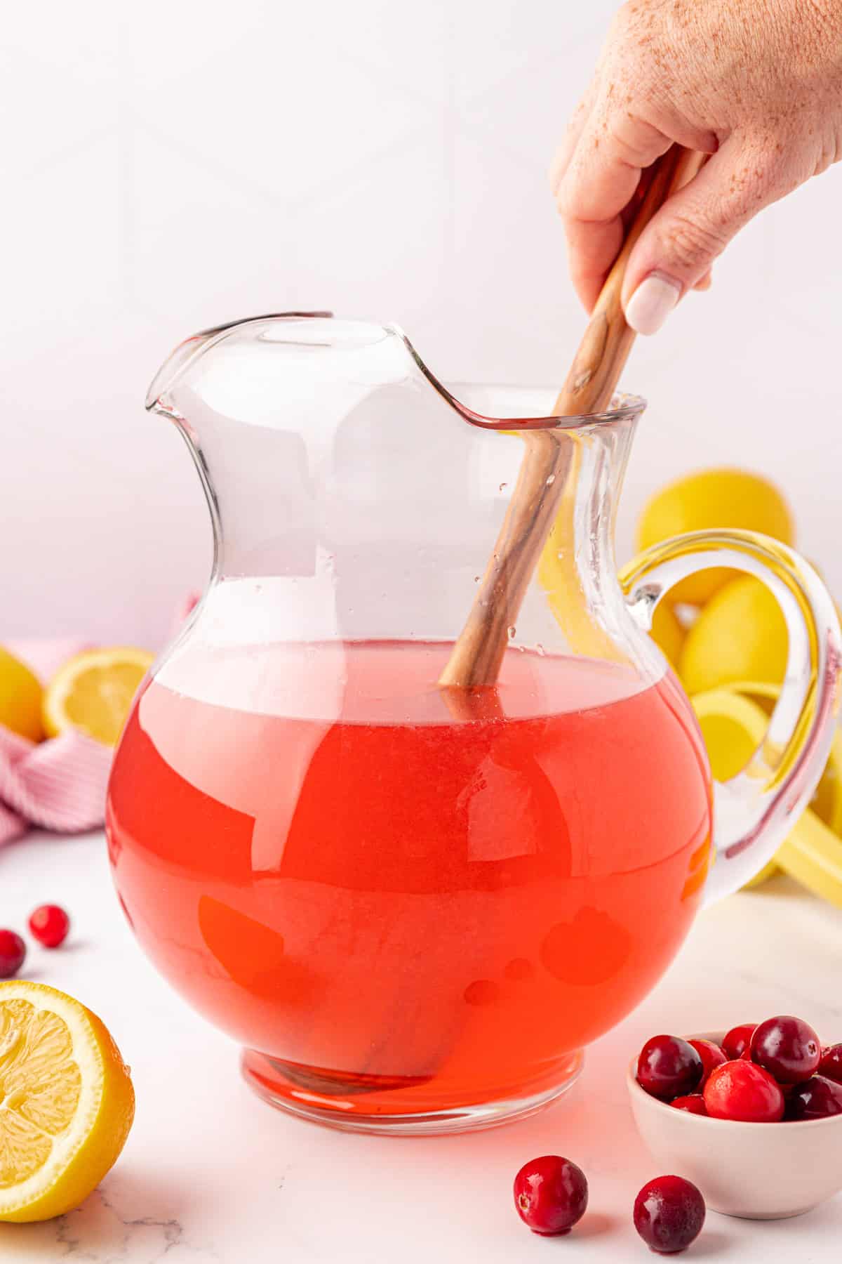 A hand stirs a pitcher of bright pink punch with a wooden spoon. The pitcher is on a white surface, surrounded by fresh lemons, cranberries, and a small bowl of cranberries.