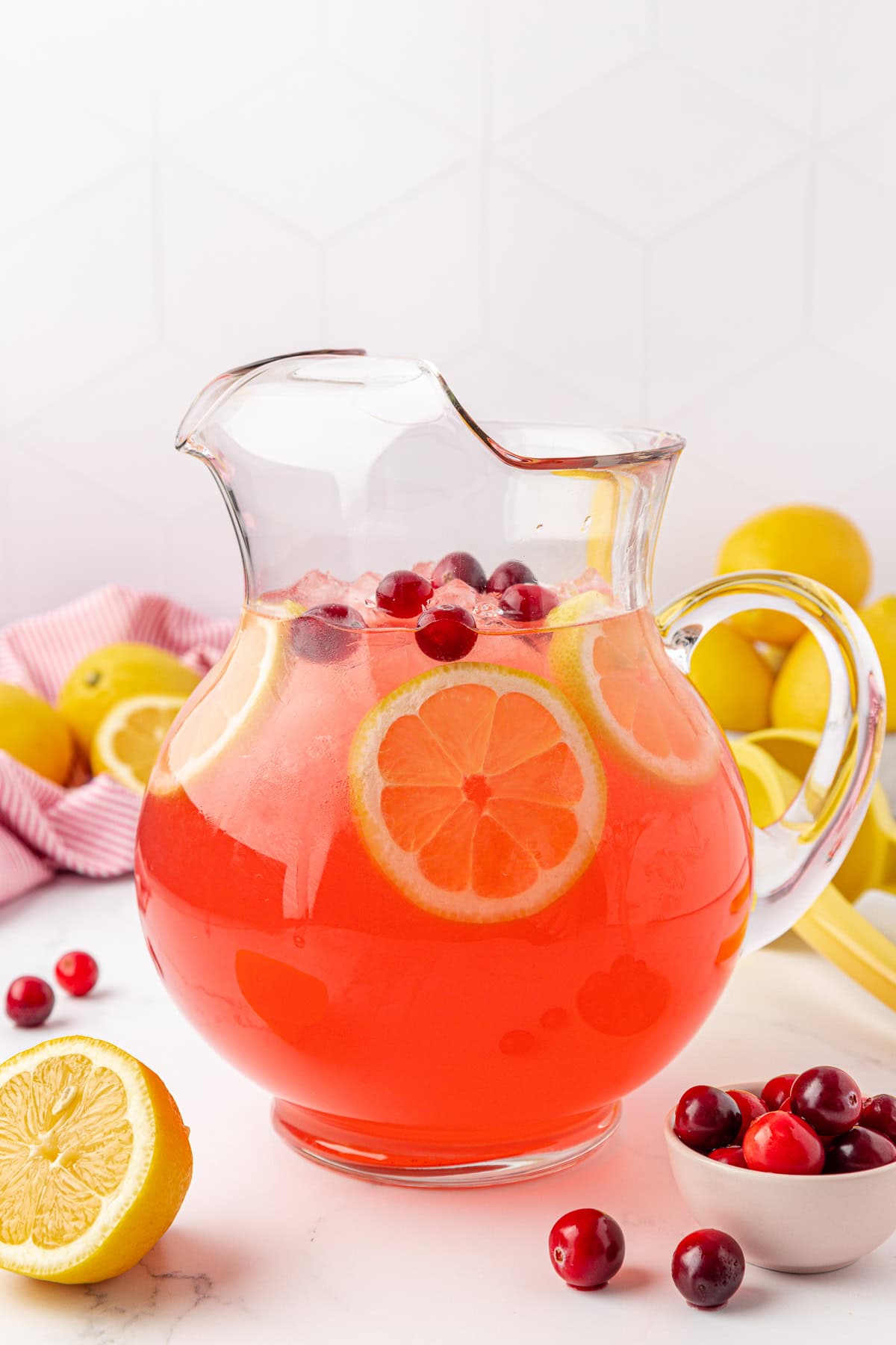 A clear glass pitcher filled with pink lemonade, garnished with lemon slices and cranberries, sits on a white counter near a small bowl of cranberries and fresh lemons.