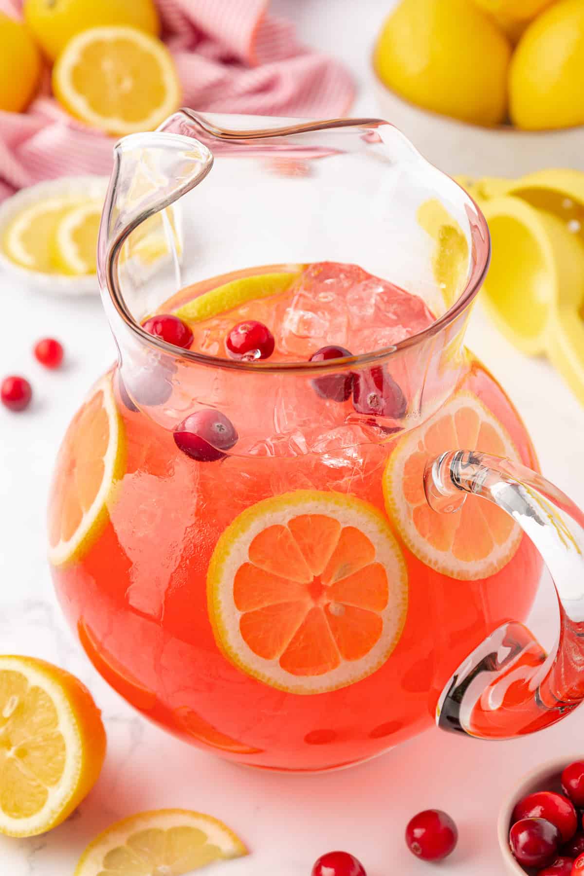 A glass pitcher filled with pink lemonade, ice cubes, lemon slices, and cranberries. Fresh lemons and a red-striped towel are in the background on a white surface.