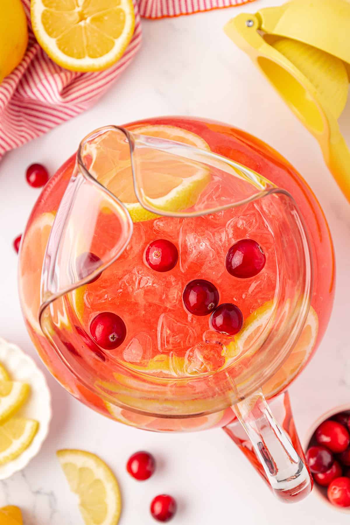 A glass pitcher filled with pink lemonade, ice, lemon slices, and cranberries sits on a white surface surrounded by fresh lemon wedges, cranberries, a citrus juicer, and a red-striped cloth.