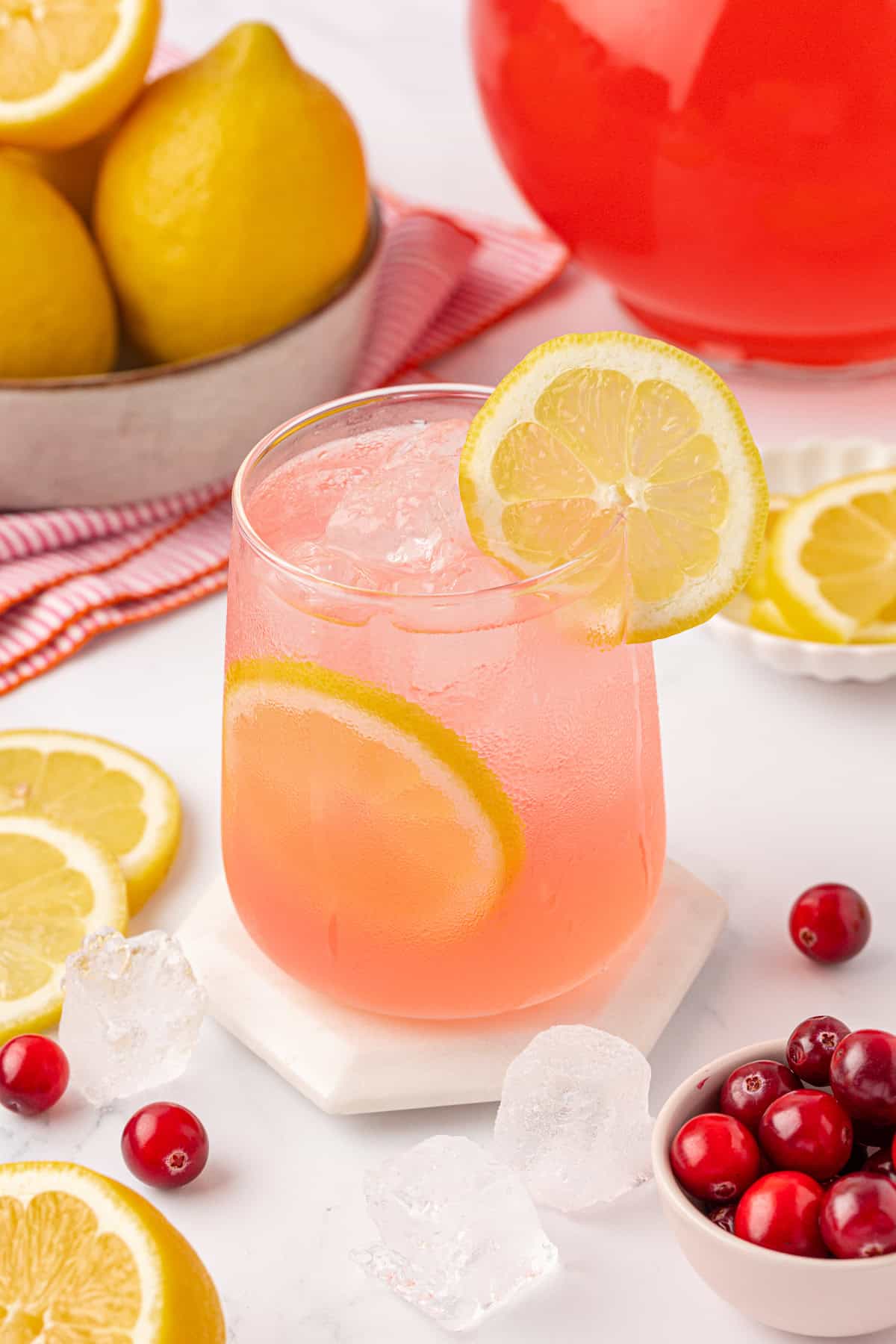 A glass of pink lemonade with ice and lemon slices, garnished with a lemon wheel, sits on a coaster. Surrounding the glass are fresh lemons, cranberries, ice cubes, and a red pitcher.
